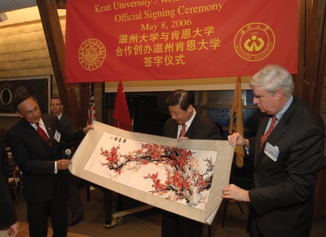 Three men in suits hold a painting under a red banner with Chinese characters on it. 